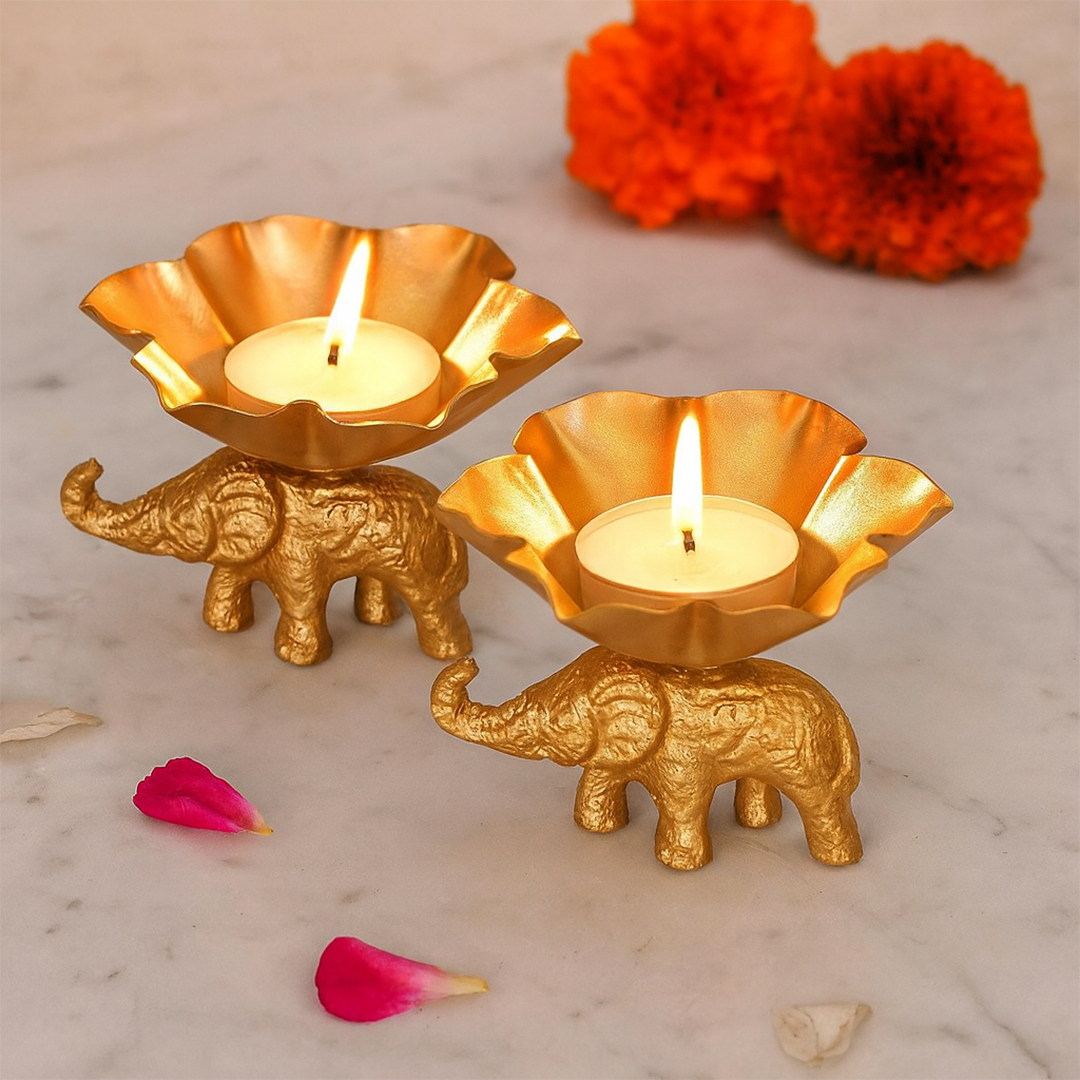 Two gold elephant candle holders with lit candles on a marble surface with red and pink flowers.